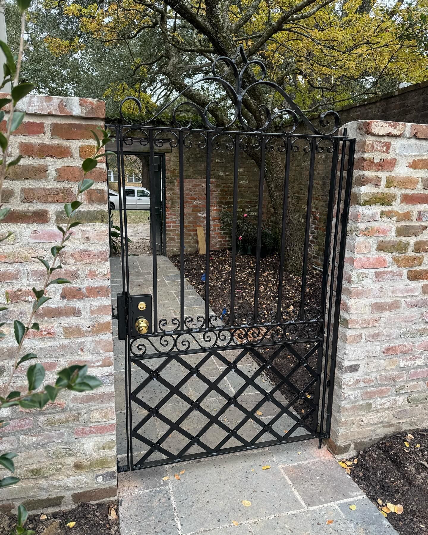 Black wrought-iron gate between brick pillars, set in a stone path with trees behind.