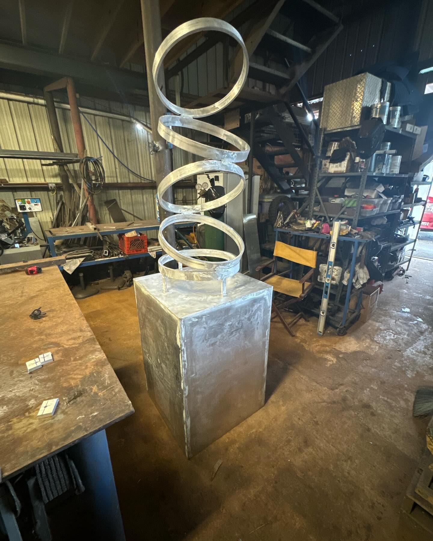 Metal spiral sculpture on a pedestal in a cluttered workshop with tools and shelves.