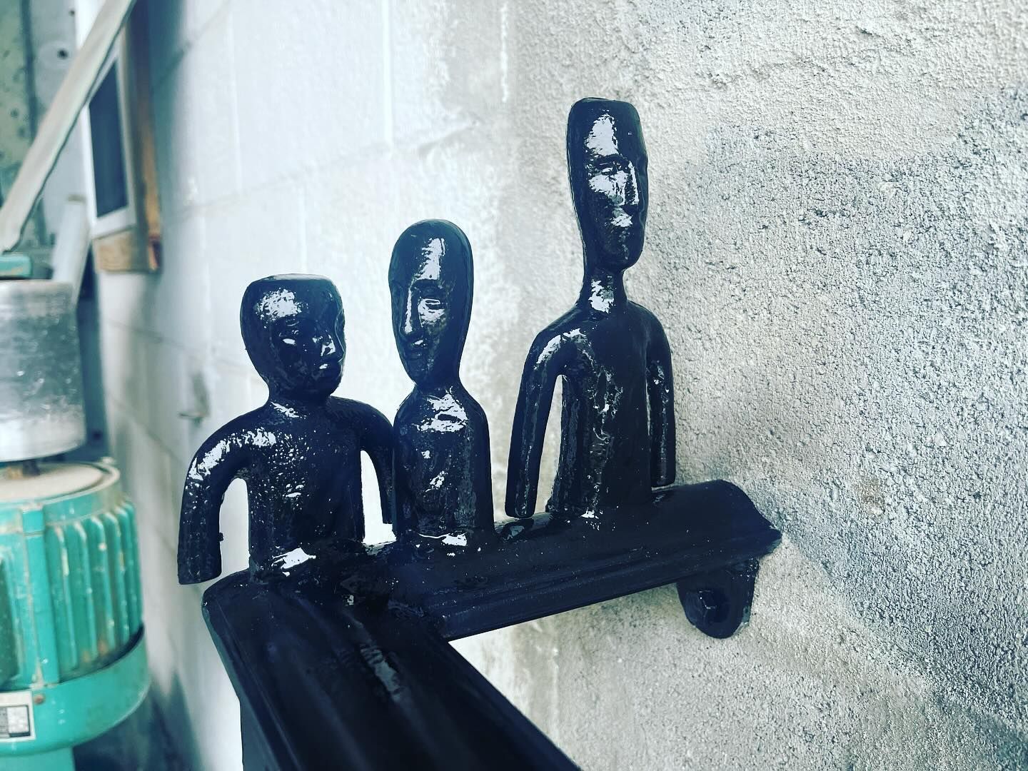 Three small black figurines seated on a wall-mounted bench against a textured wall.