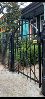 Black iron gate in front of a house with teal and blue siding and greenery along a gravel path