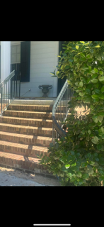 Brick steps leading up to a house porch with white siding, metal railings, and green bushes beside it