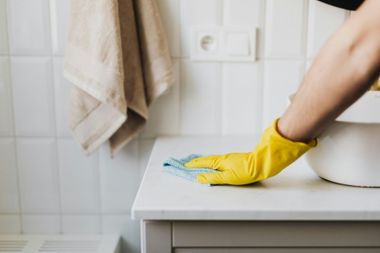 A person wearing a yellow glove cleans a white bathroom counter with a blue cloth.