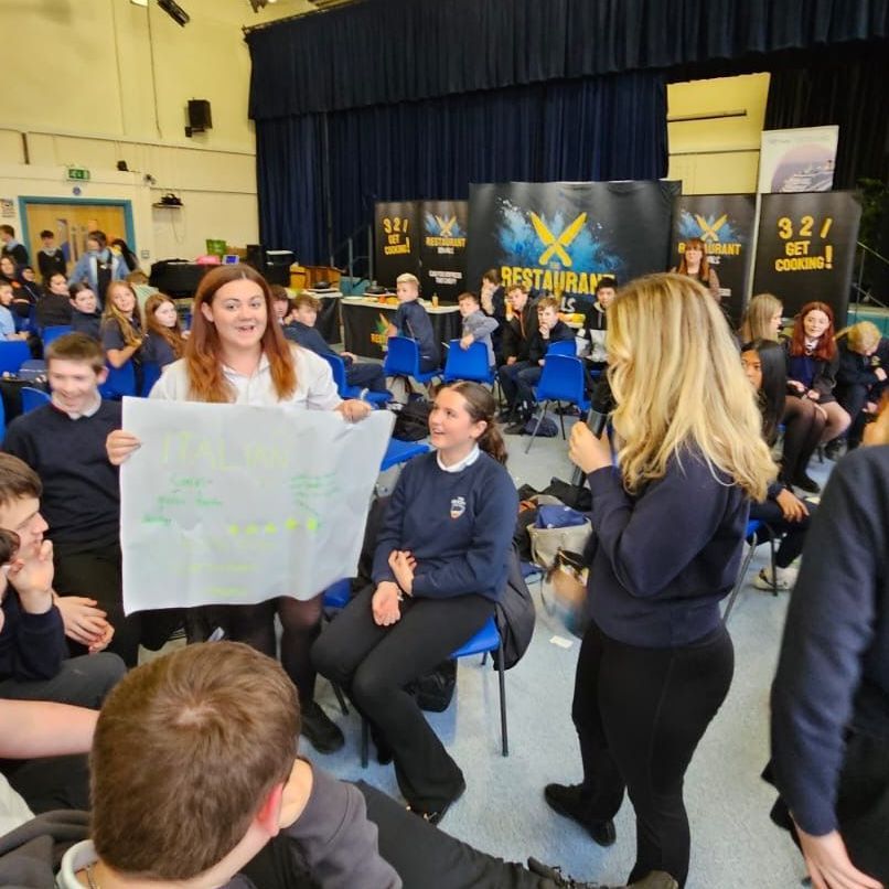 Students in a school hall brainstorm, holding papers. A woman smiles and presents a sheet. Other students watch.