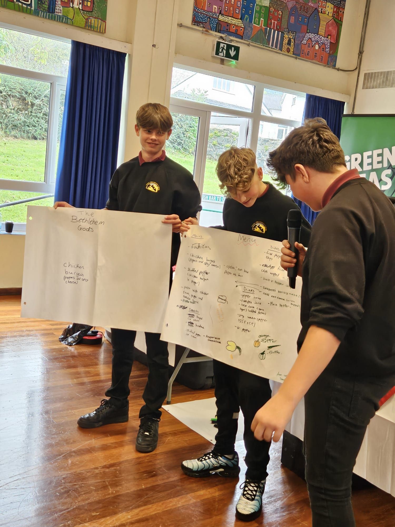 Three students presenting posters at an event. They are standing indoors with posters showing text and diagrams.