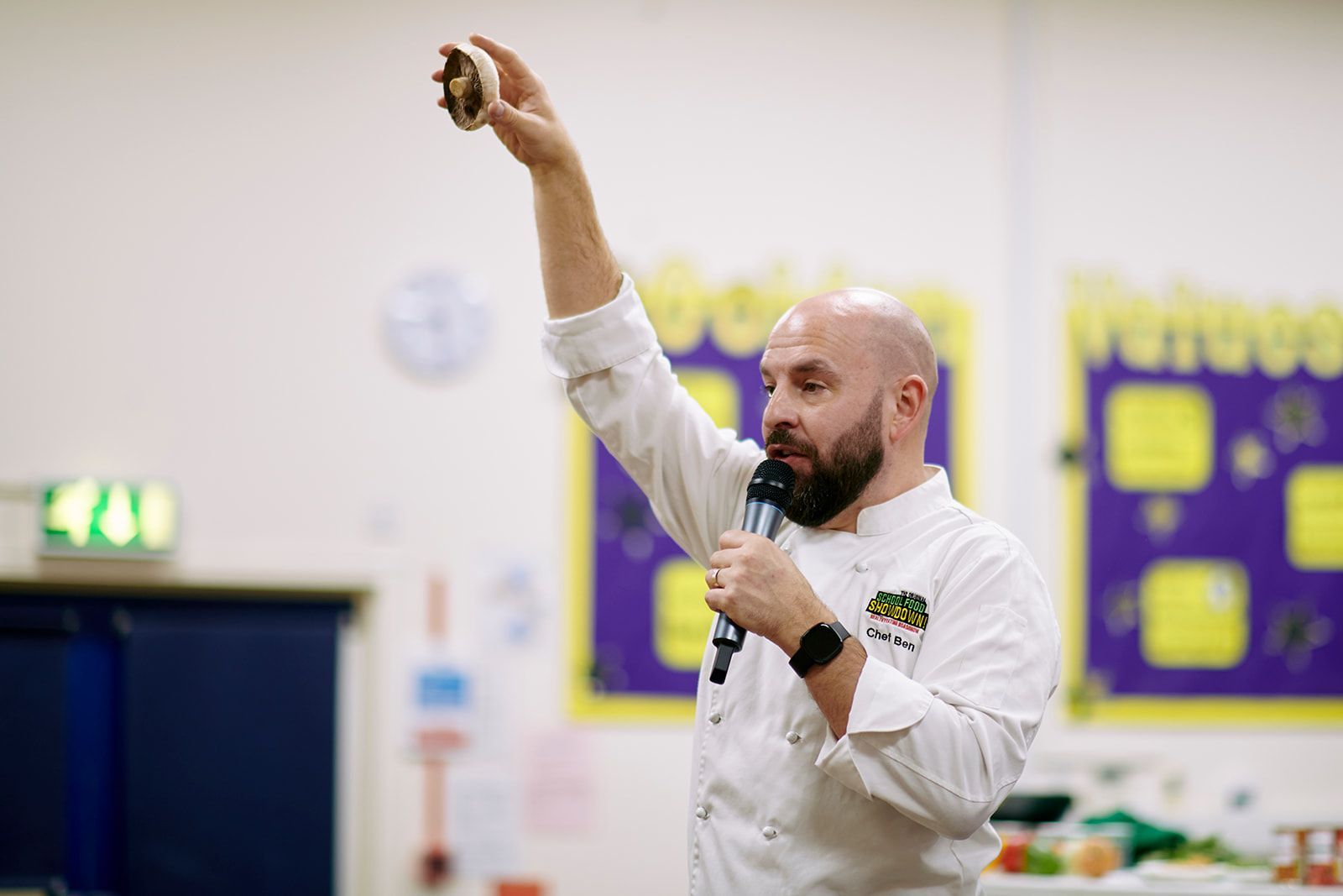 Chef holding baked good, speaking into a microphone with raised arm in a school setting.
