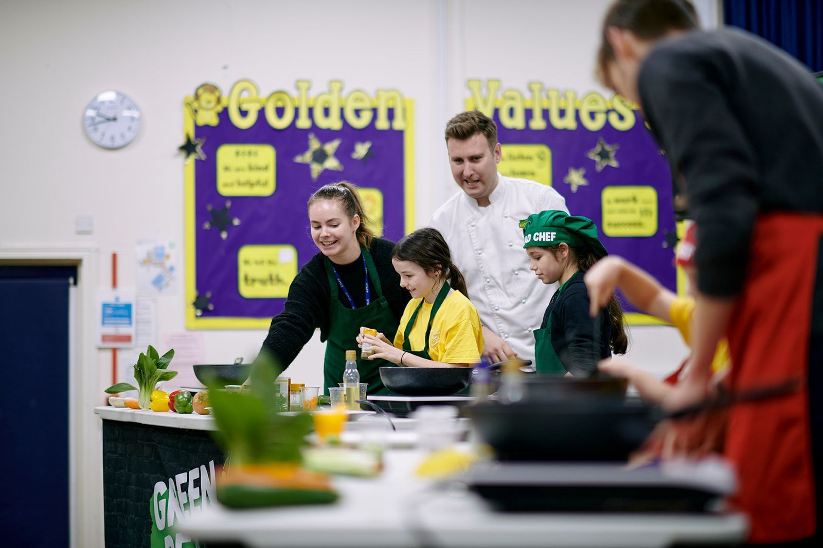 People cooking, likely in a school kitchen. A chef assists children in cooking class.