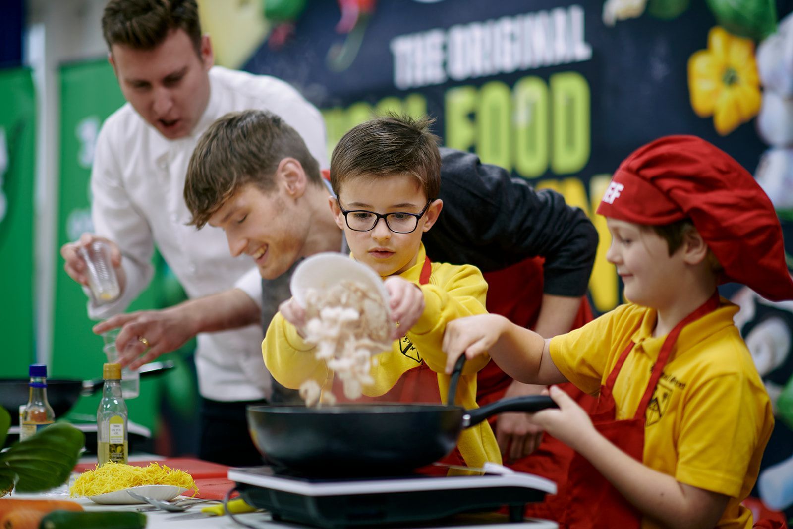 Children and adults cooking together at a food event. They're adding ingredients to a pan, wearing aprons and chef hats.