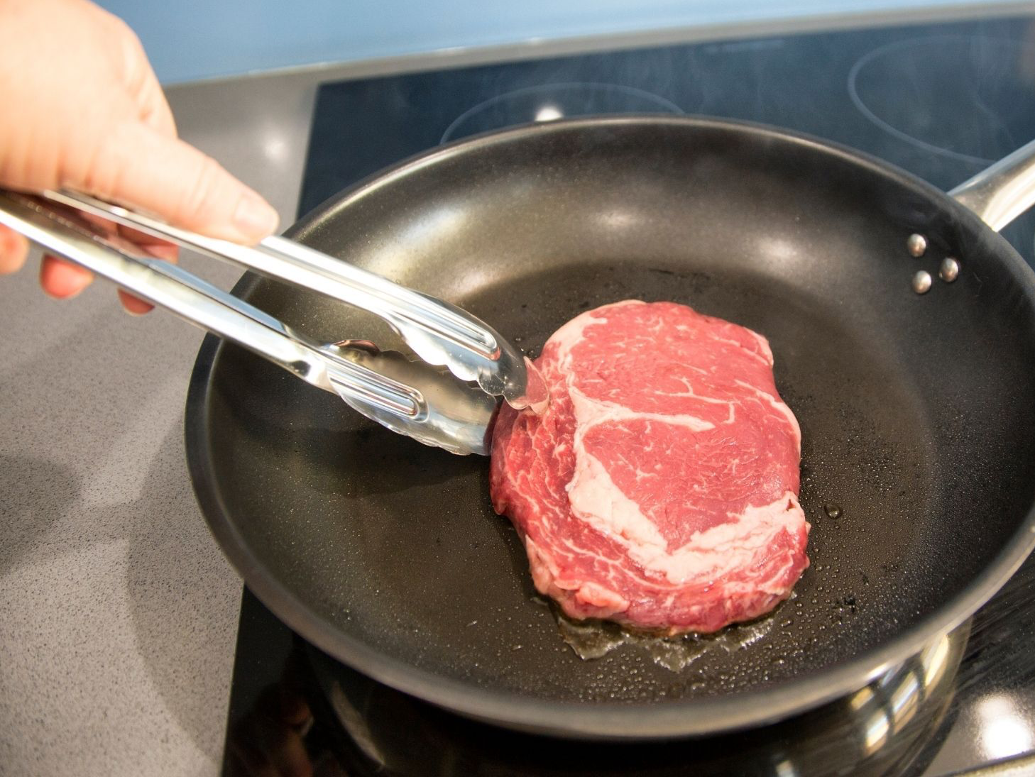 Hand using tongs to flip a raw steak in a hot skillet on a stovetop.