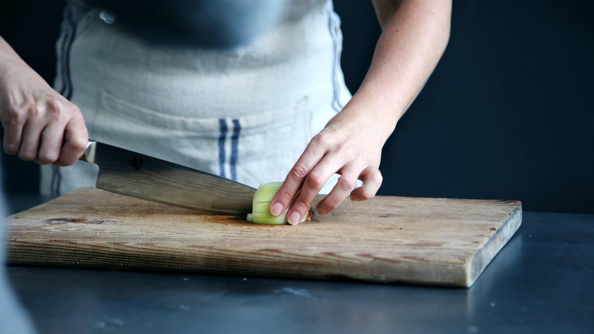 Person slicing a vegetable on a wooden cutting board with a large knife, in a kitchen.