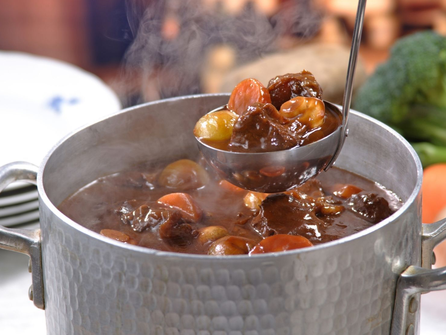 Steaming pot of beef stew being ladled out, with carrots, potatoes, and gravy.
