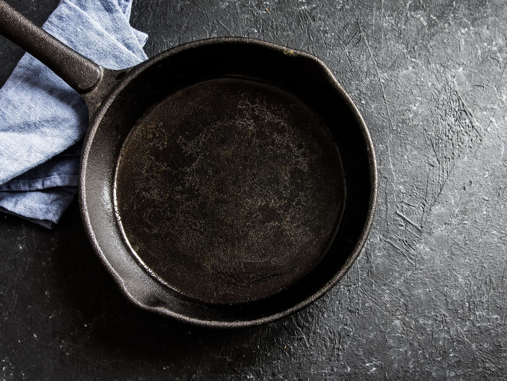 Empty, black cast iron skillet on a dark surface with a blue cloth in the background.