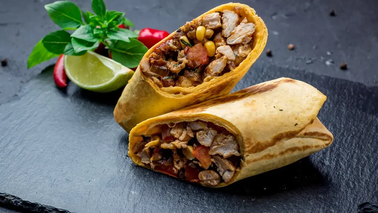 Two burritos cut in half, filled with meat, vegetables, and corn. Resting on a dark slate.