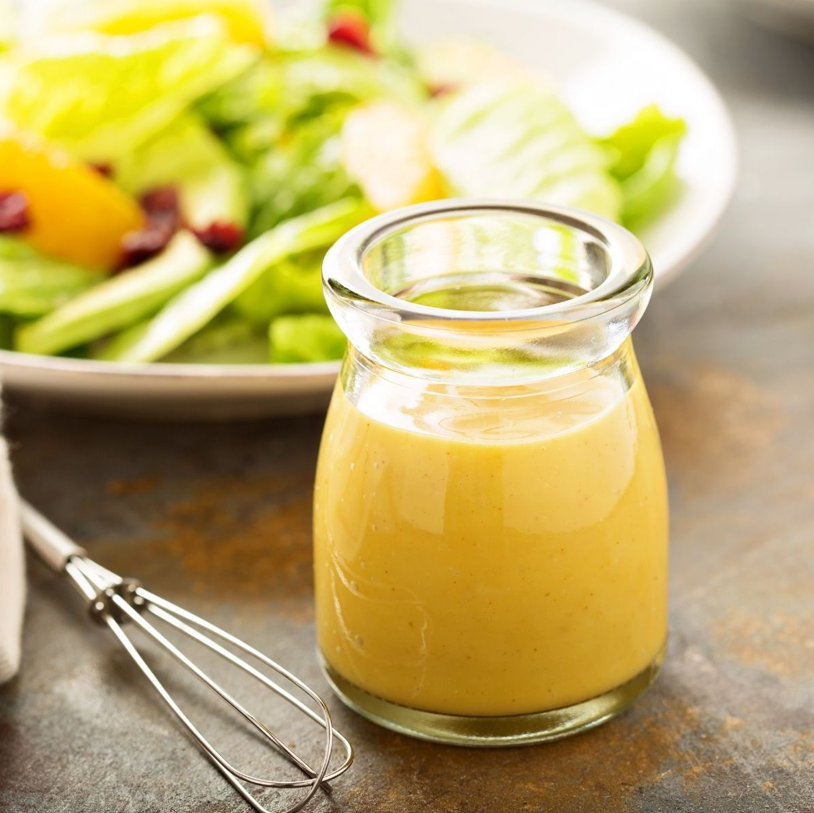Small glass jar of yellow salad dressing, with a salad in the background and a whisk beside it.