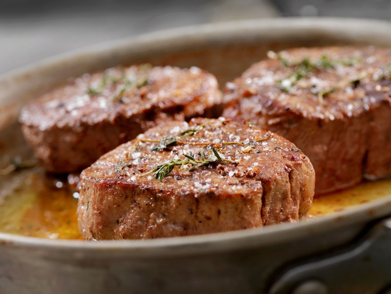 Three browned, seared steaks in a pan, sprinkled with herbs and salt, surrounded by cooking juices.