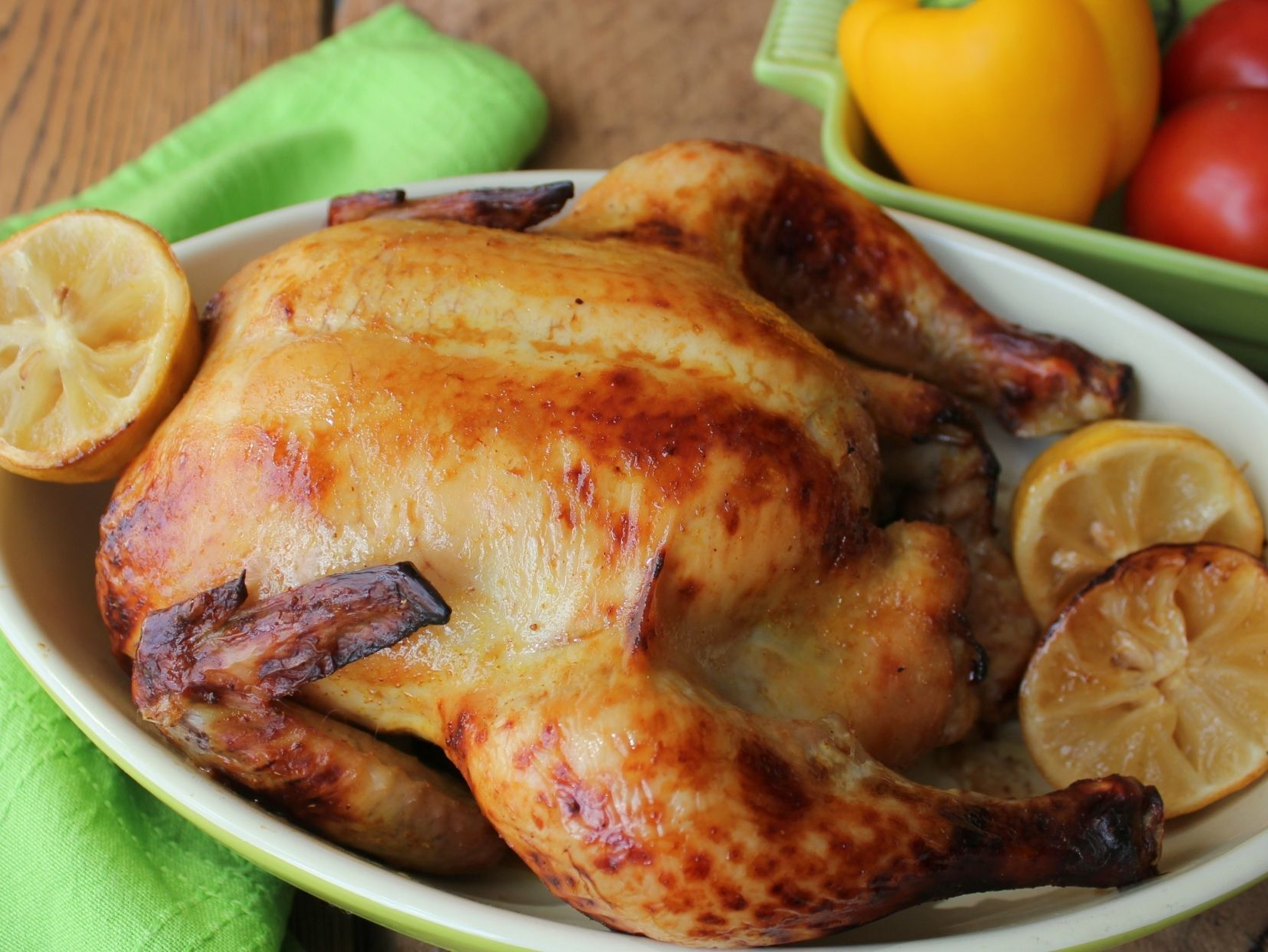 Roasted whole chicken with lemon slices in a green dish, wooden background.
