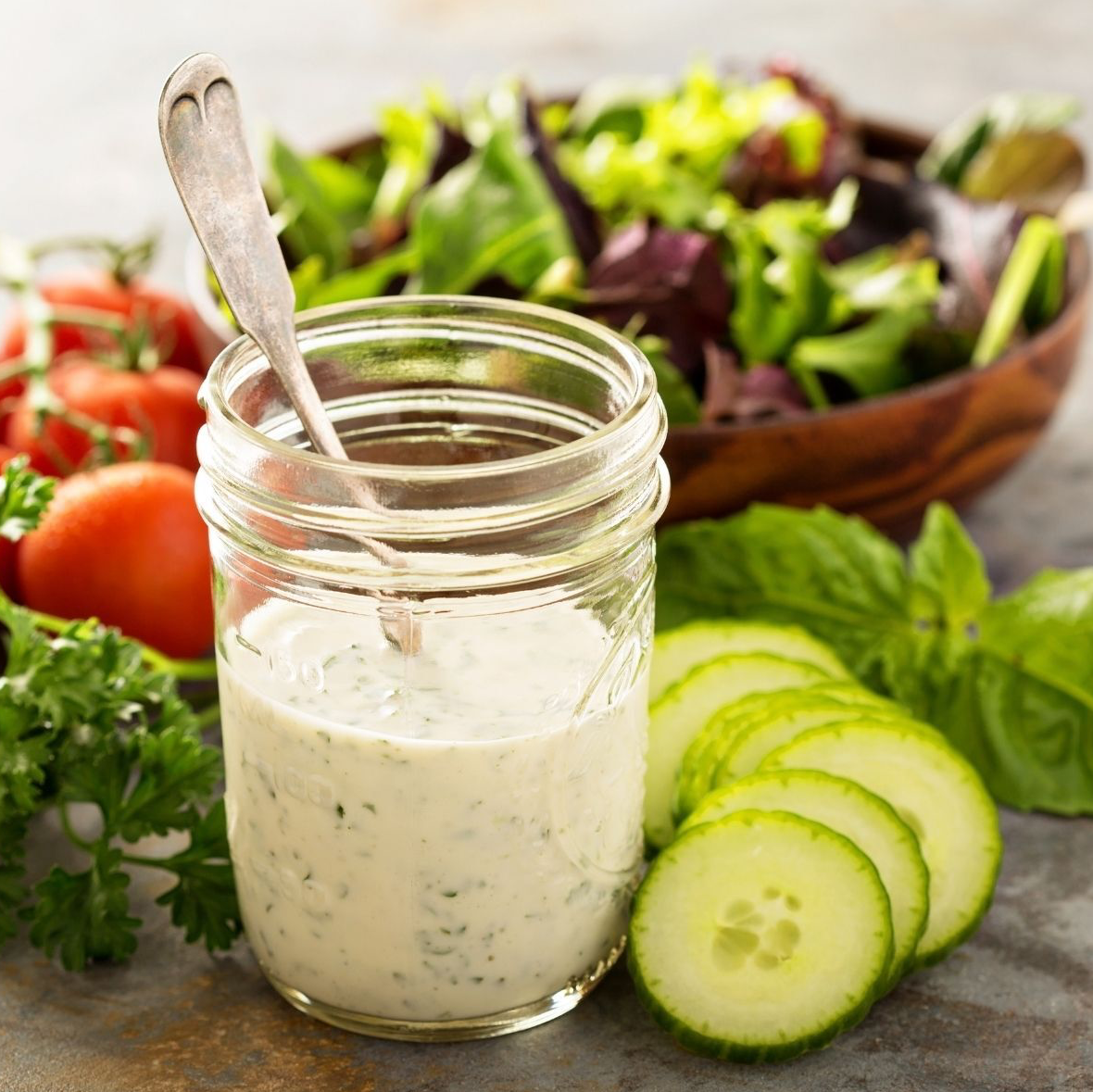 Jar of creamy salad dressing with a spoon, alongside salad, tomatoes, cucumbers, and basil.
