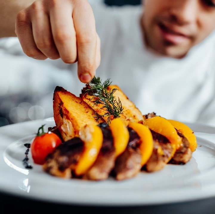 Chef garnishing a plate of food with fresh herbs. Potatoes, tomatoes, and peaches are also present.