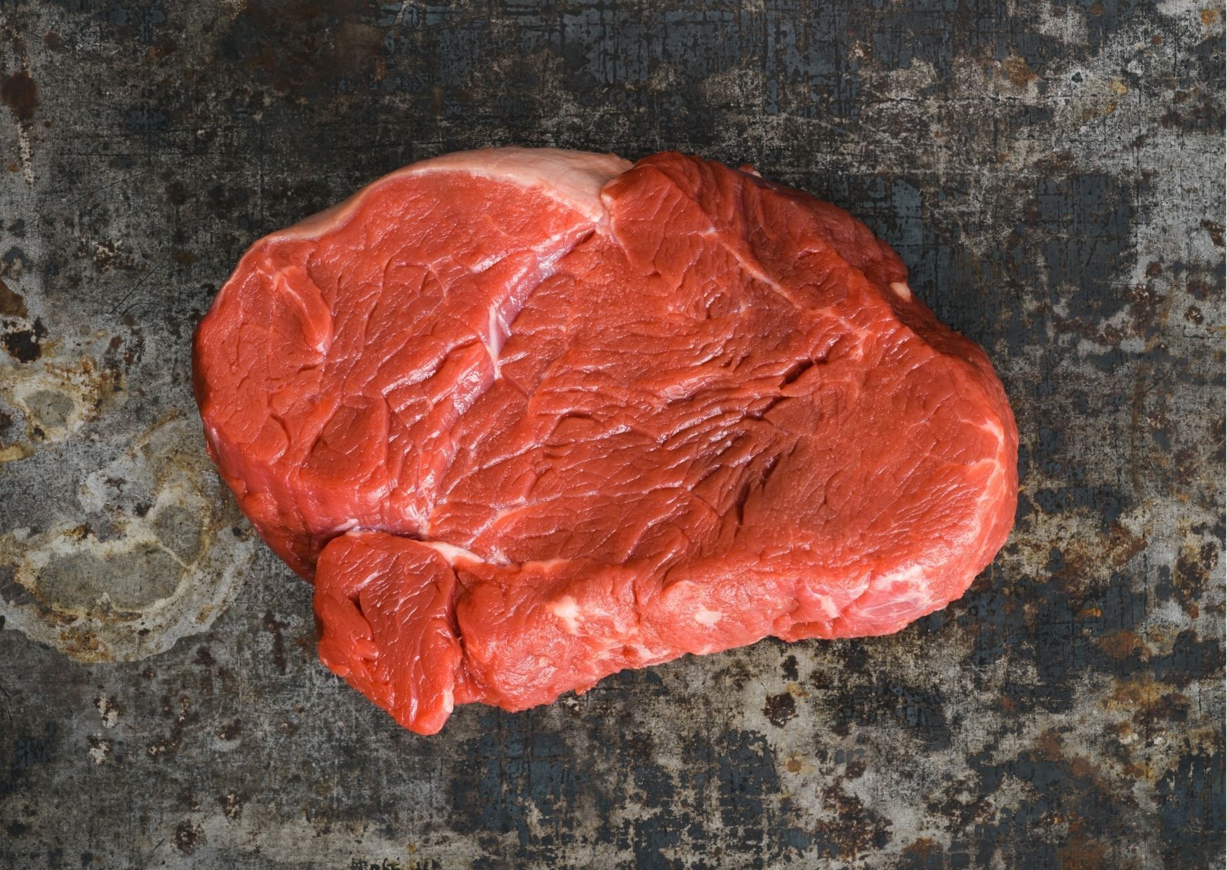 Raw, round steak with visible marbling and fat on a textured, dark, metal surface.