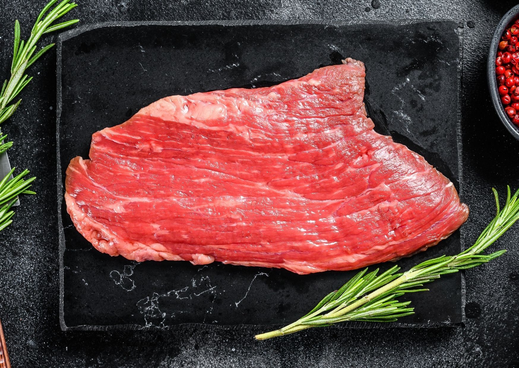 Raw, flat piece of beef on a black slate, with rosemary sprigs and peppercorns.