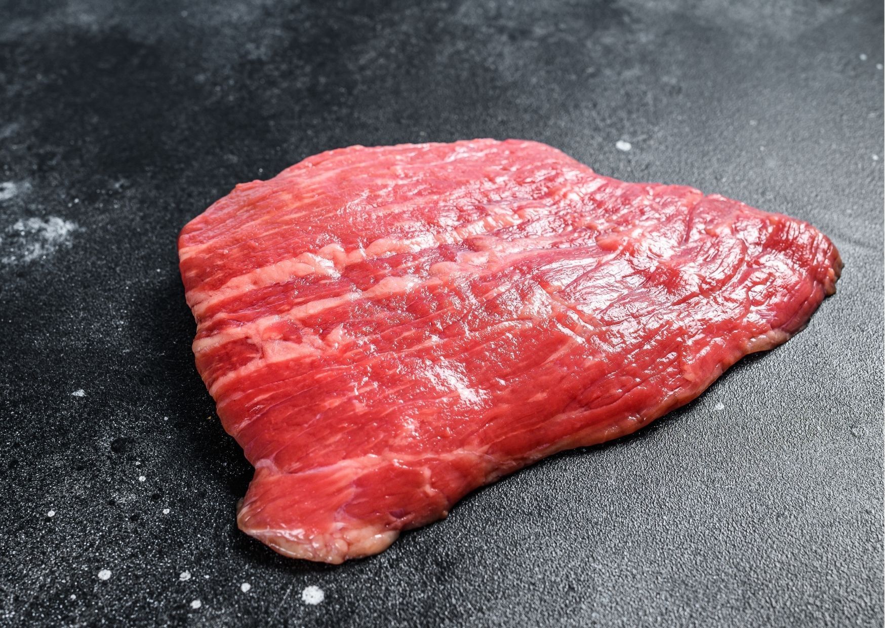 Raw, red steak with fat marbling, resting on a dark surface.