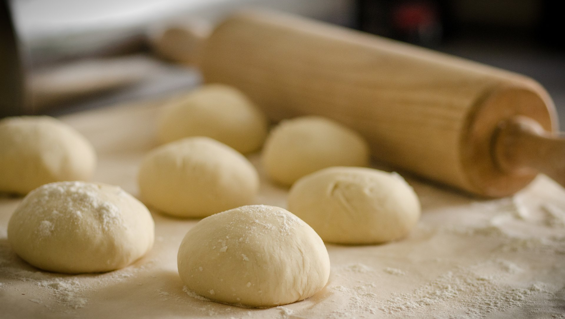 Dough balls sprinkled with flour on a wooden surface, with a rolling pin in the background.