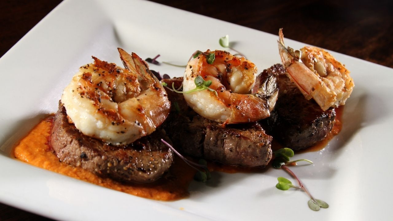 Seared steak topped with shrimp on a white plate, with orange sauce.