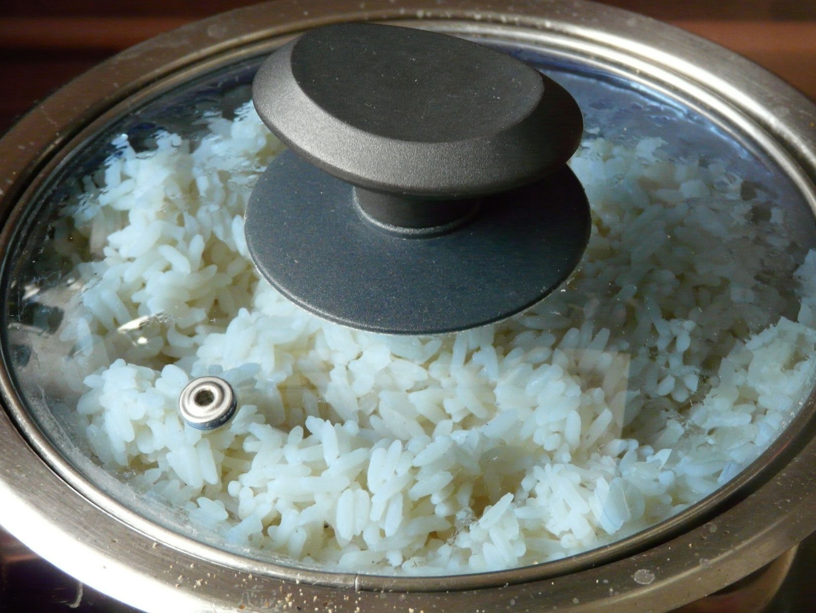 Cooked white rice in a pot, partially covered with a glass lid.