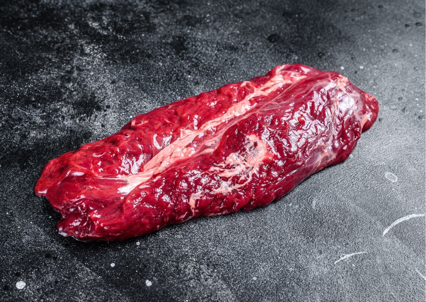 Raw, red flank steak on a dark, textured surface.