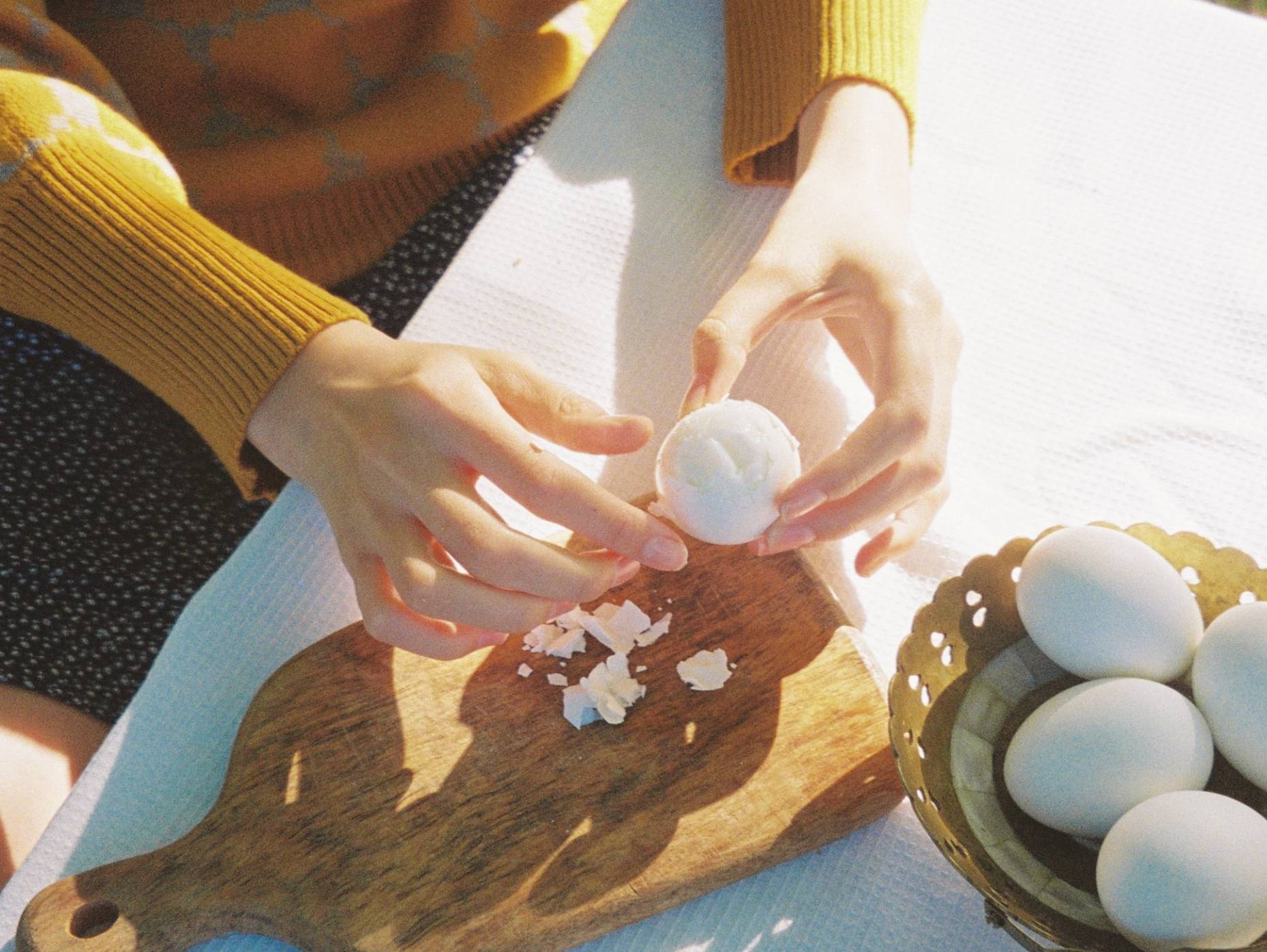 Hands peeling a hard-boiled egg on a wooden board. A basket of eggs sits nearby.