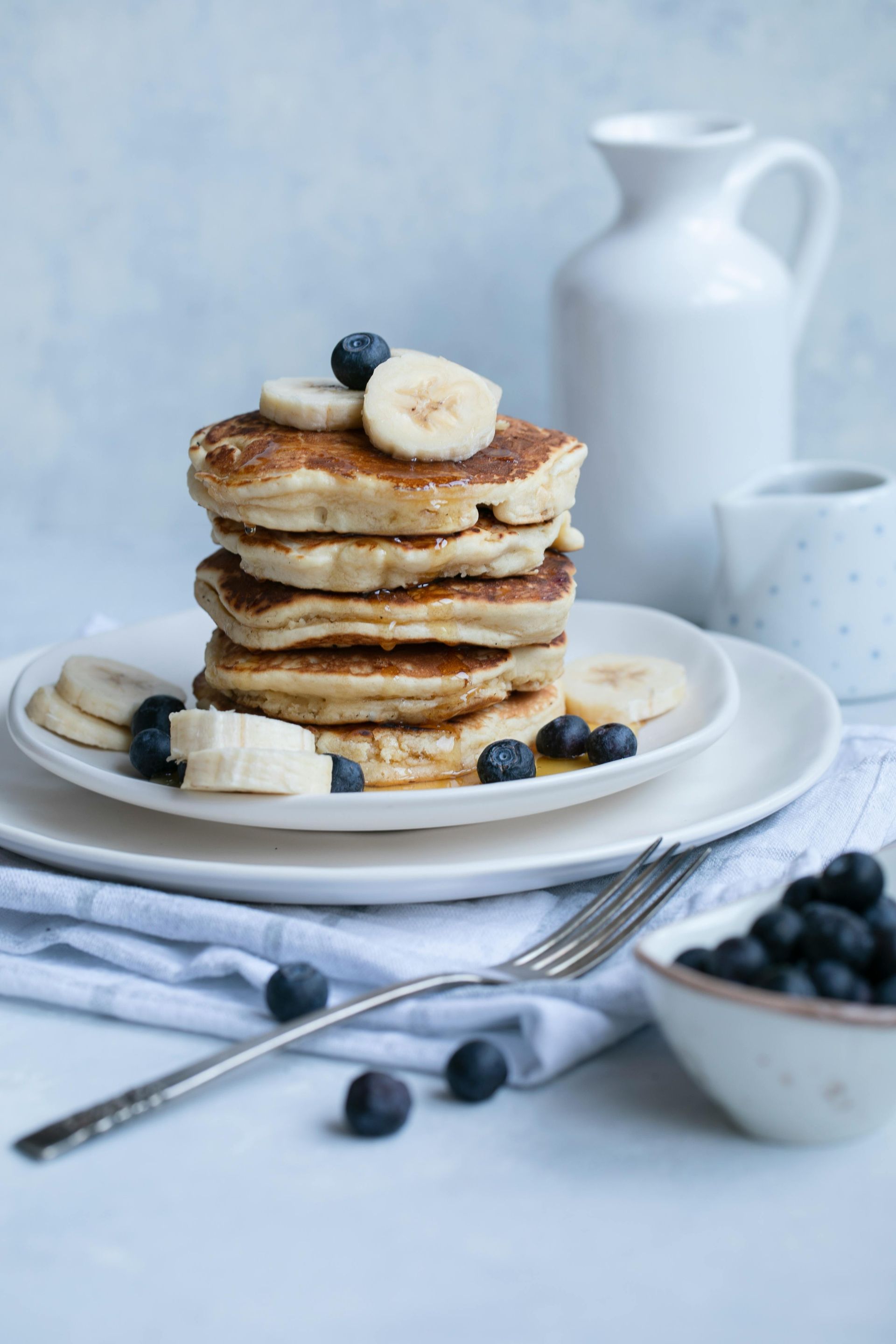 Stack of pancakes with banana slices and blueberries on white plates.
