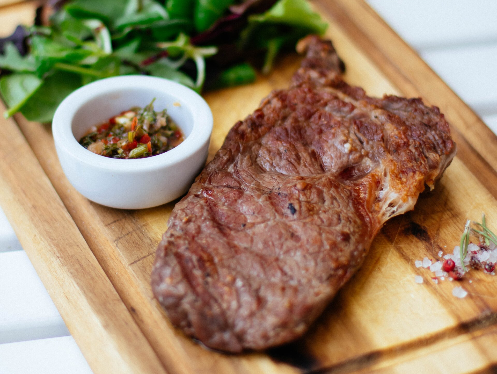 Grilled steak with sauce, greens, and salt on a wooden board.