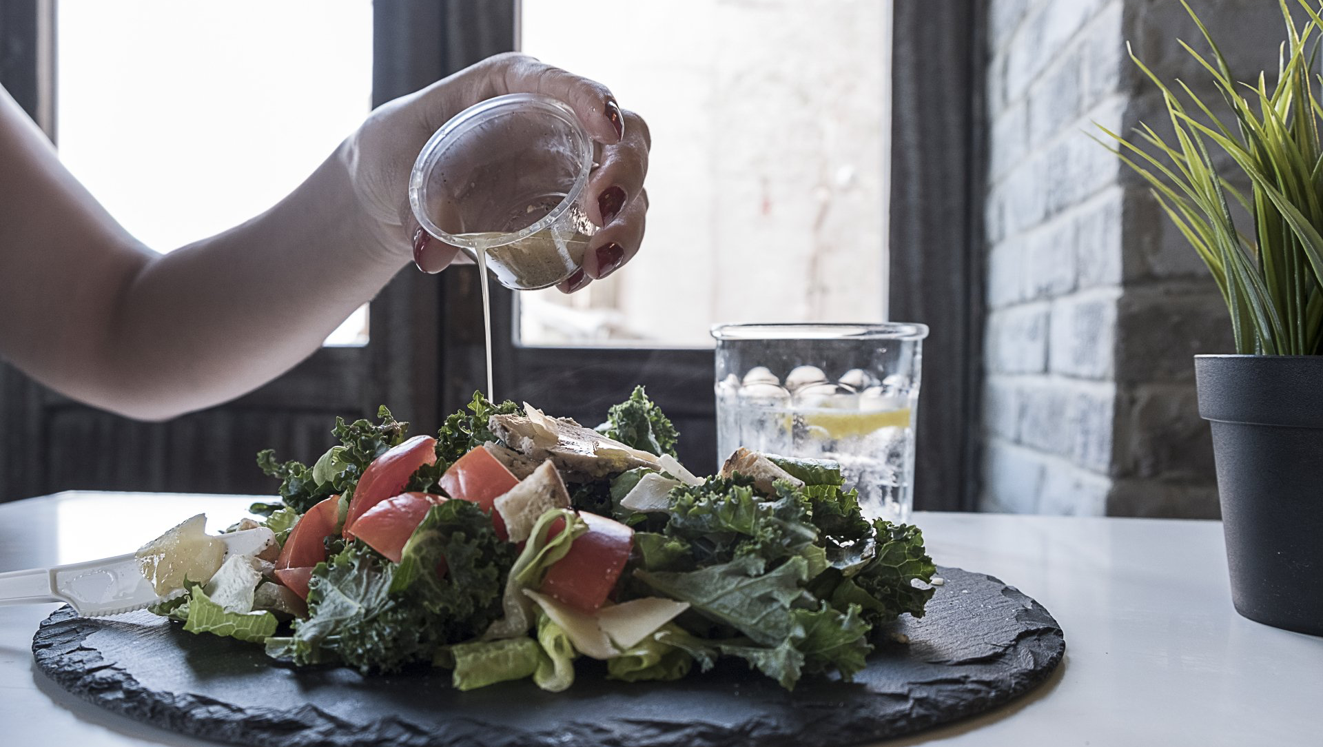 Person squeezing lemon juice onto a salad on a slate plate. Water glass and plant visible.