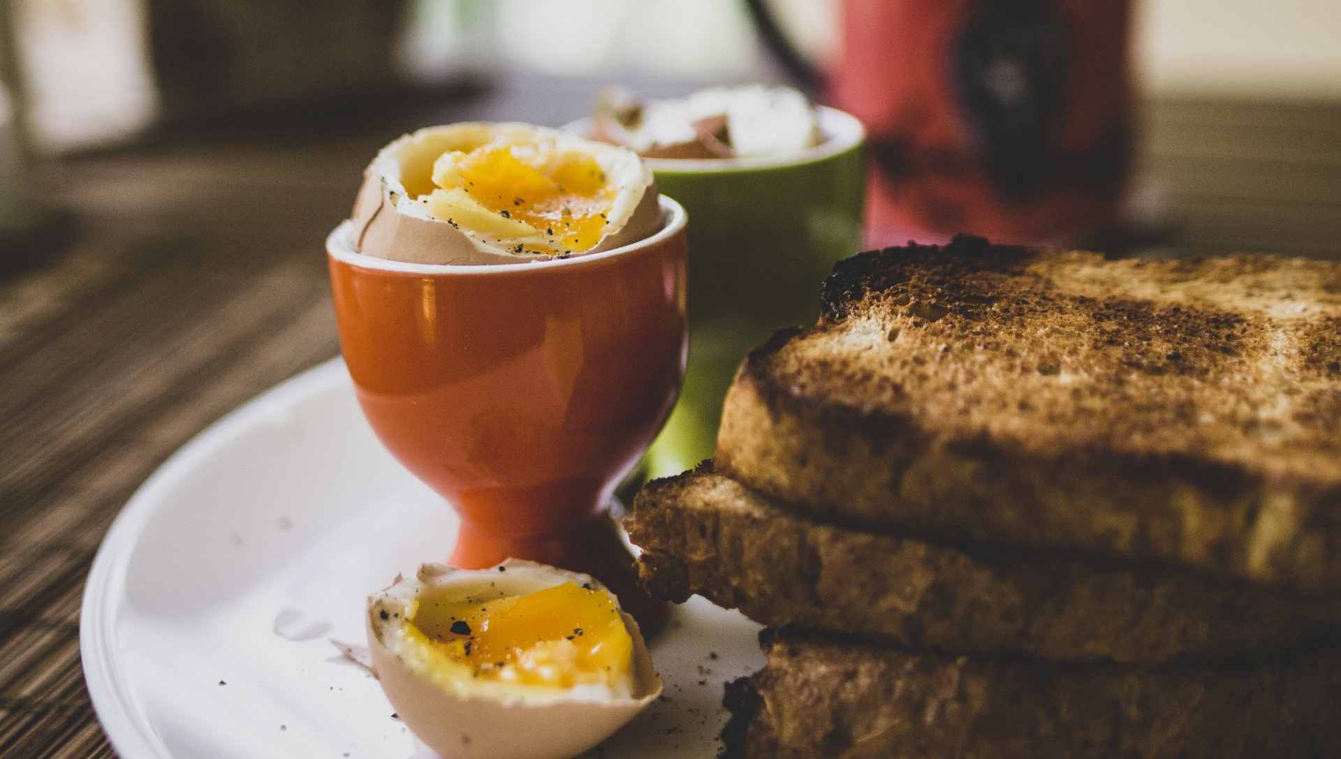 Soft-boiled eggs in orange and green cups, with toast, on a white plate.