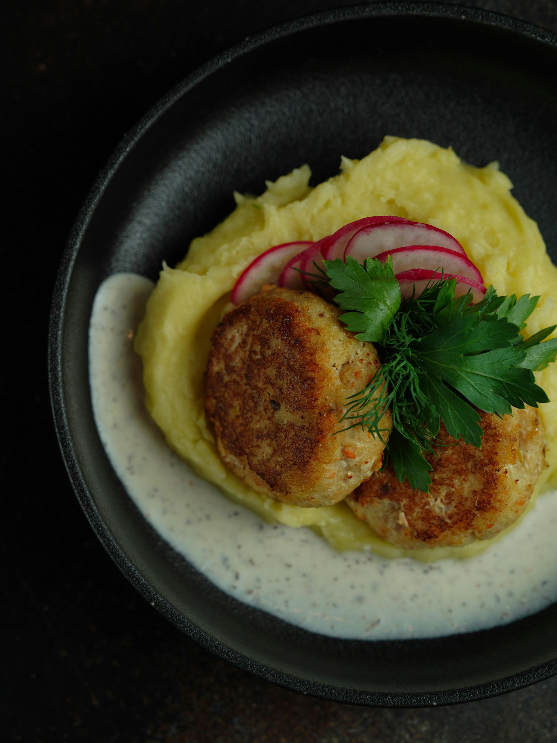 Two golden patties on creamy mashed potatoes, topped with parsley and radish slices in a dark bowl