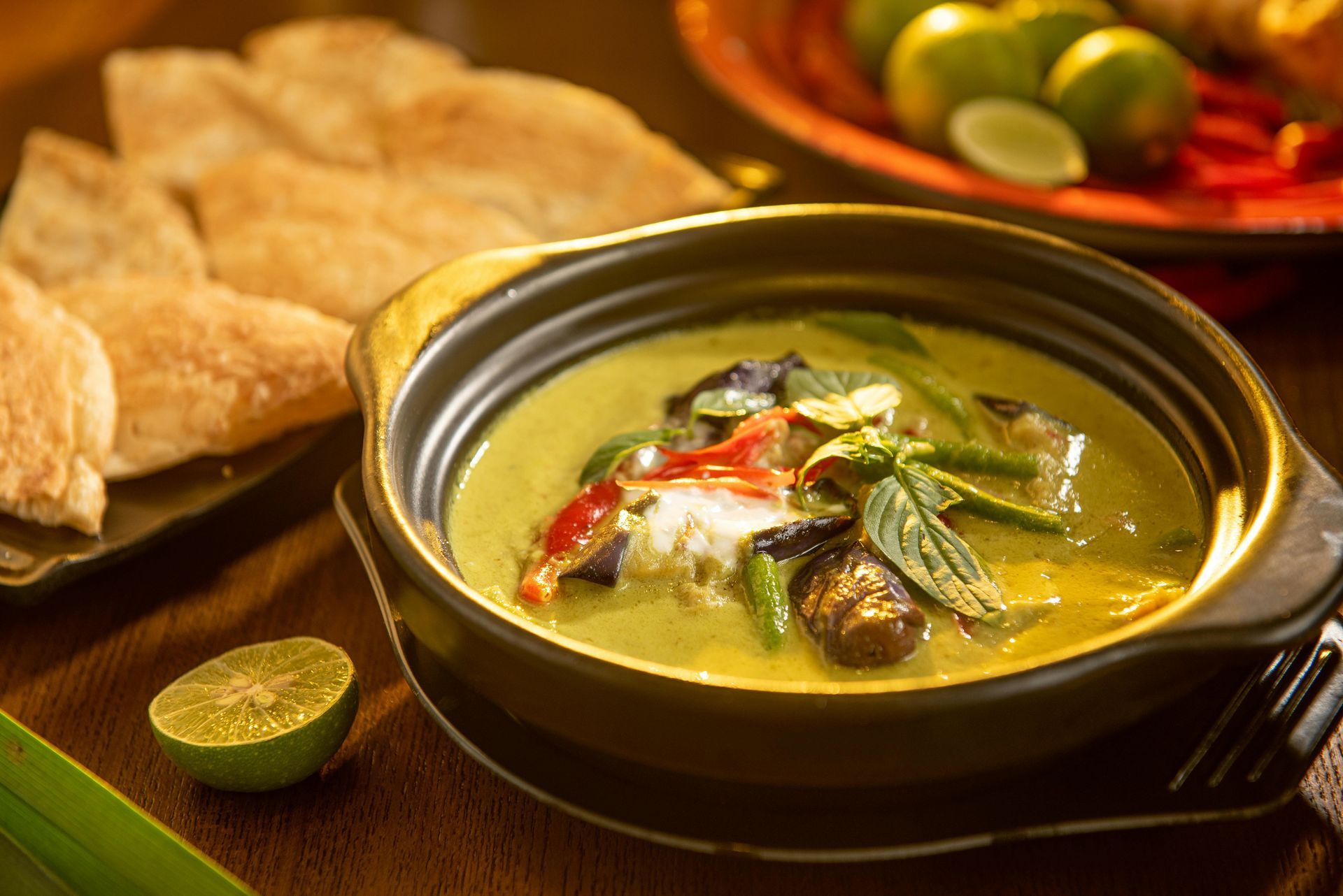 Hot curry in a black bowl with lime, red chilies, and bread on a wooden table