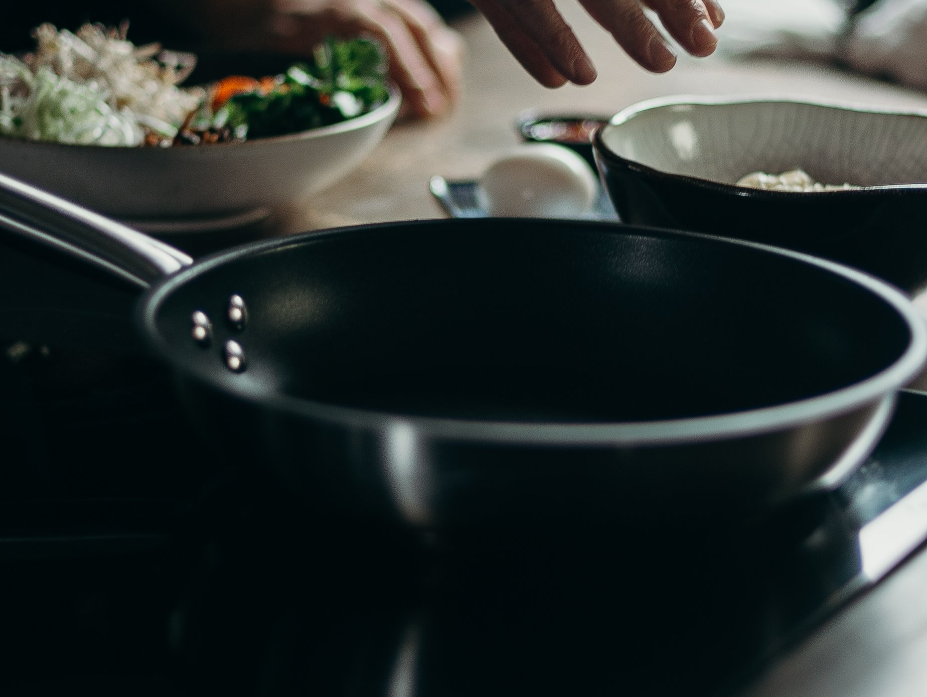 Empty black frying pan on a stovetop with bowls of ingredients in the background, a hand reaching over the pan.