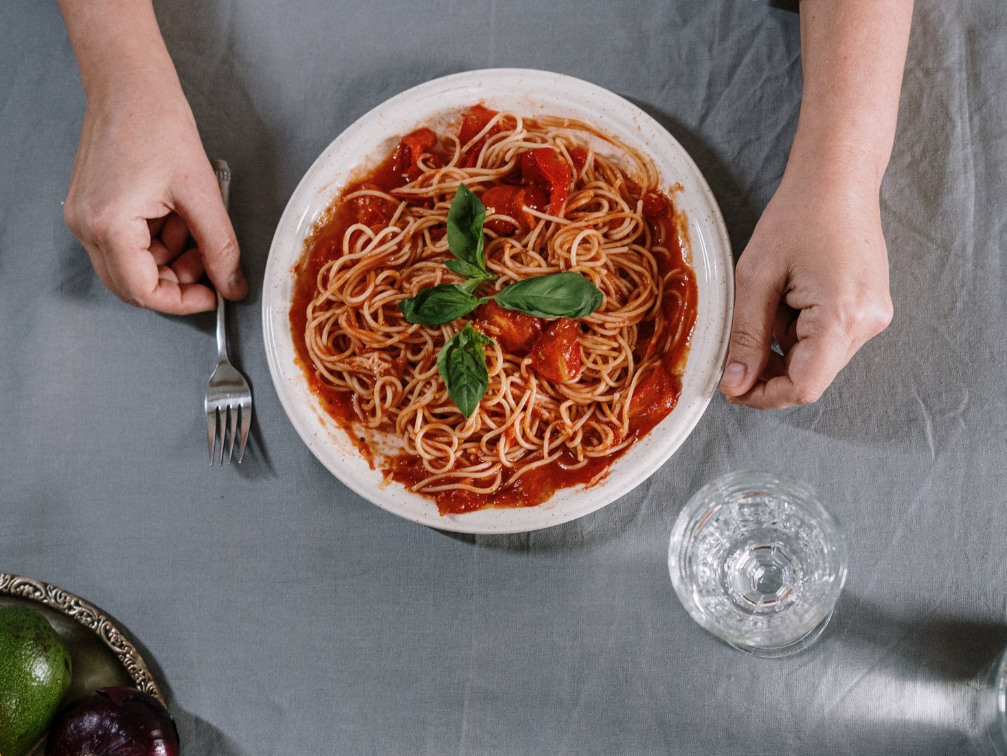 Hands holding a plate of spaghetti with tomato sauce, basil, and a glass of water on a gray tablecloth.