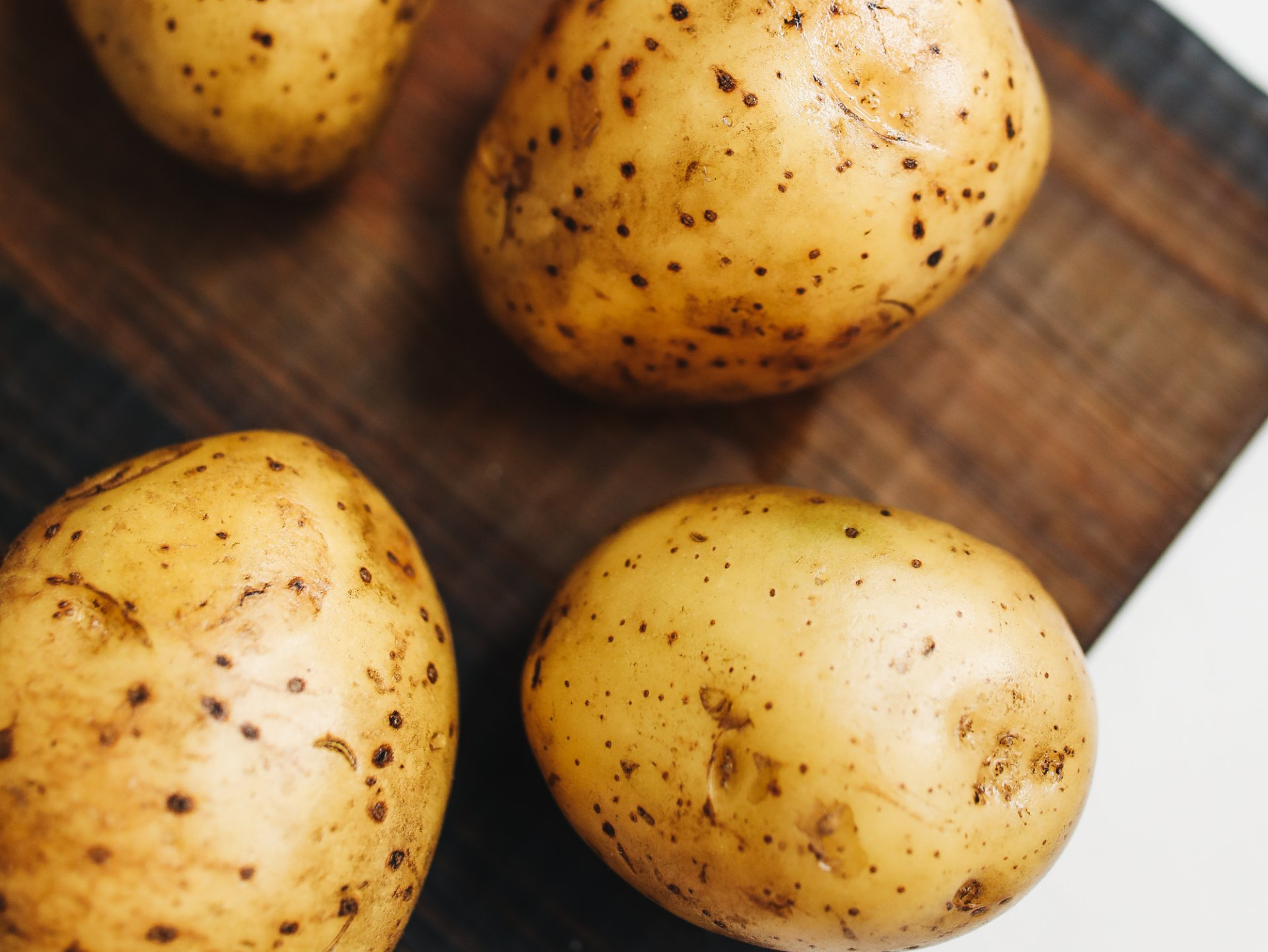 Four unpeeled potatoes on a dark wooden cutting board, with speckled, light-brown skin.