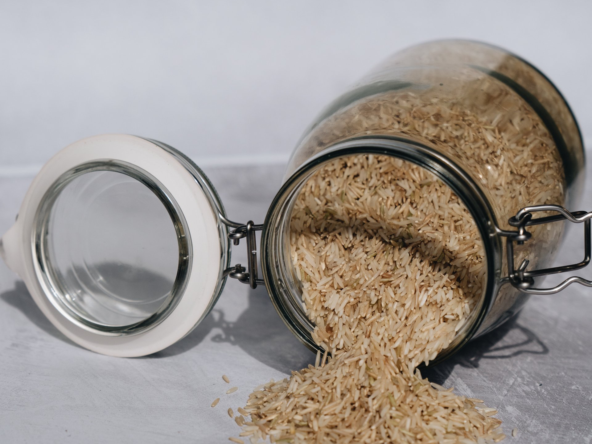 Glass jar of brown rice spilling onto a grey surface; lid is open.