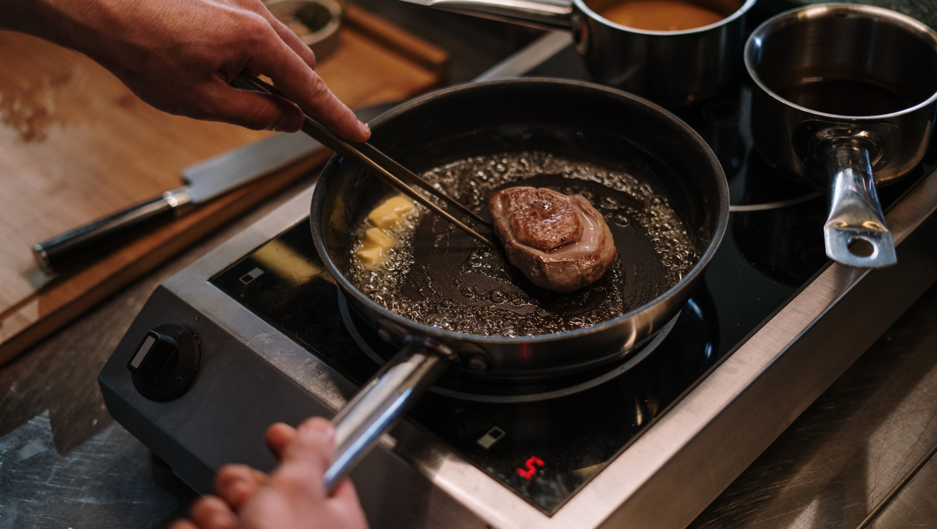 Person using tongs to flip a steak in a pan on a stovetop, with butter and sauce nearby.