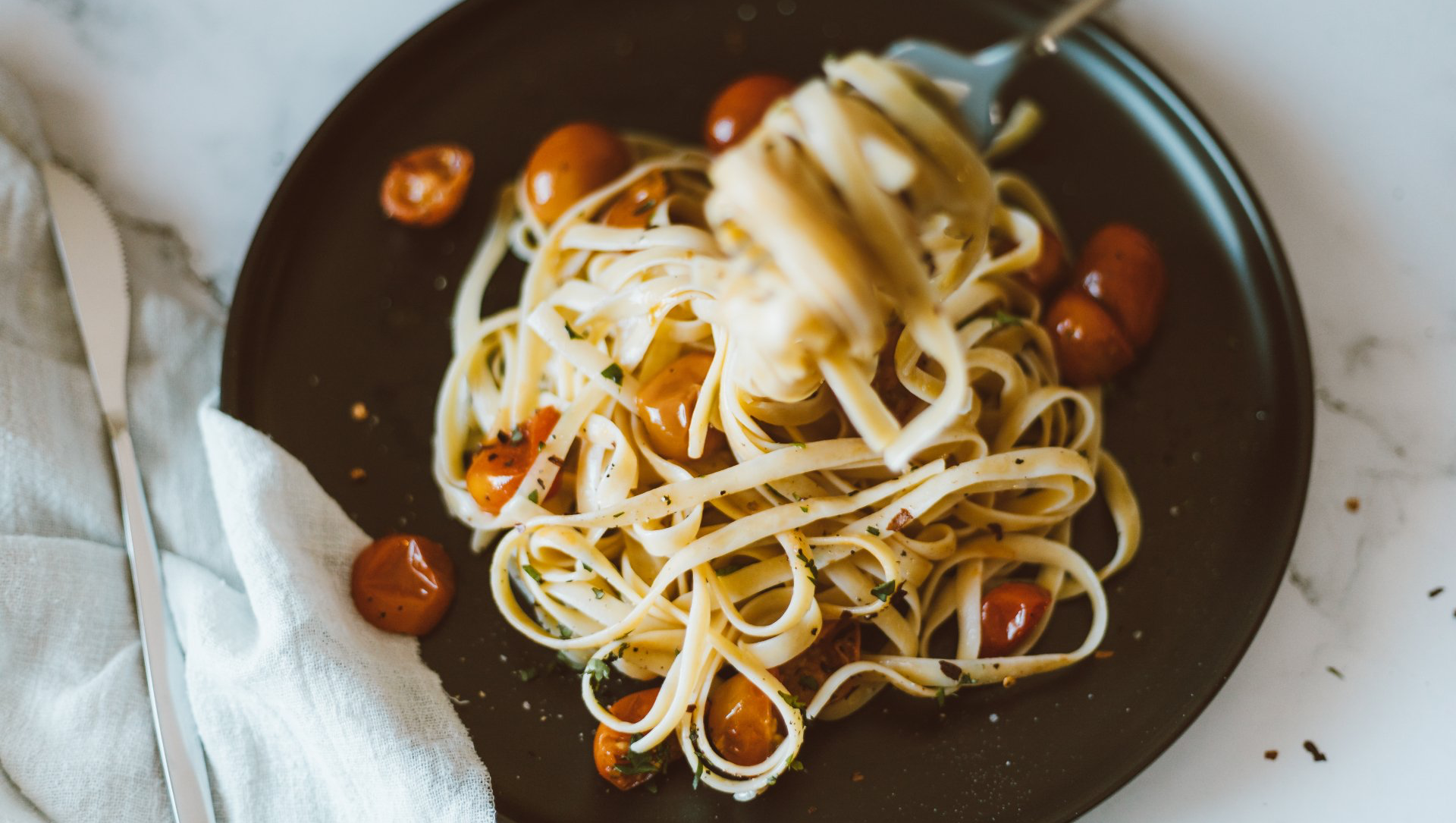 Pasta with cherry tomatoes on a dark plate, fork lifting a portion, beside a knife and napkin on a marble surface.