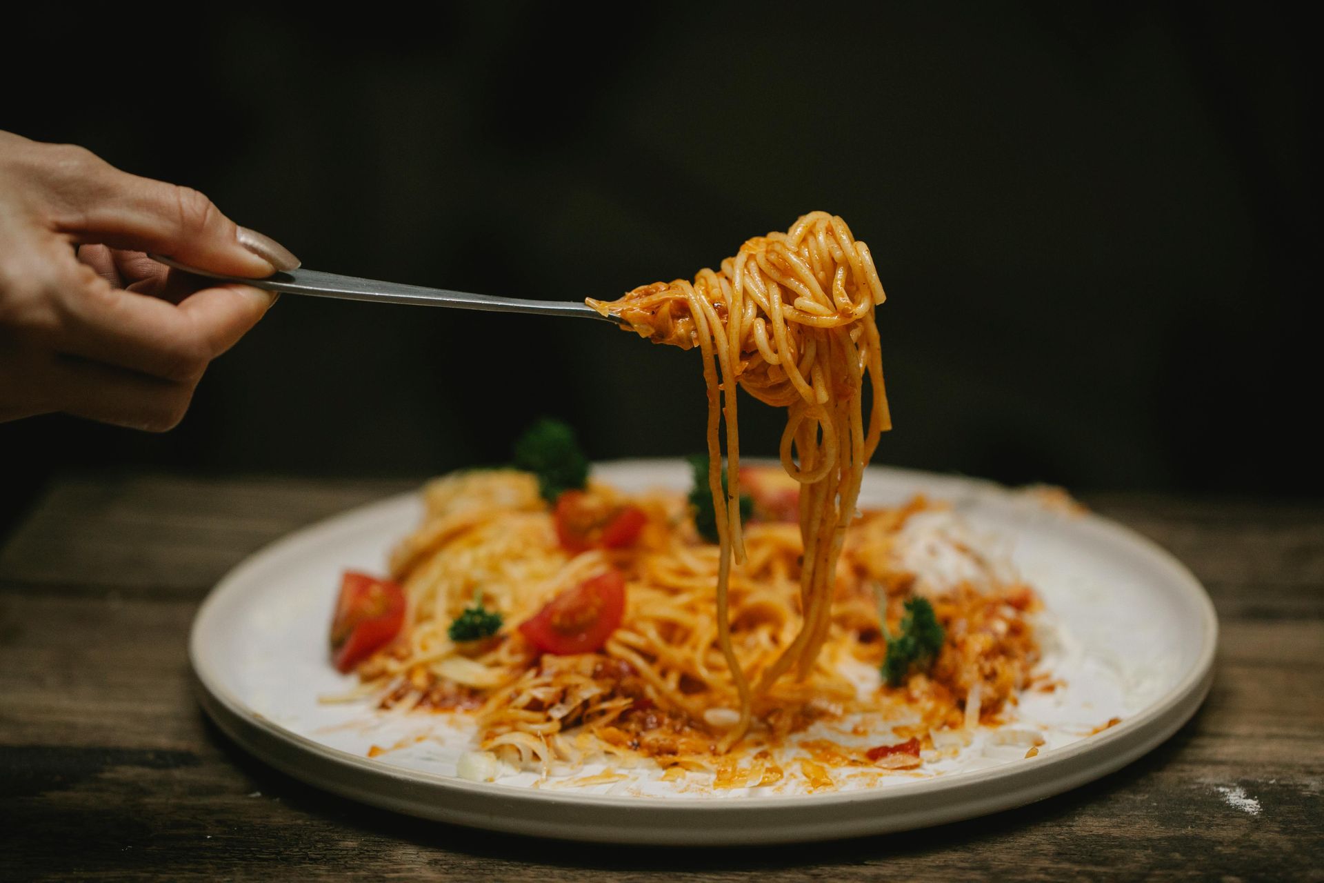 Hand holding fork with spaghetti over a plate of pasta with tomatoes and greens.