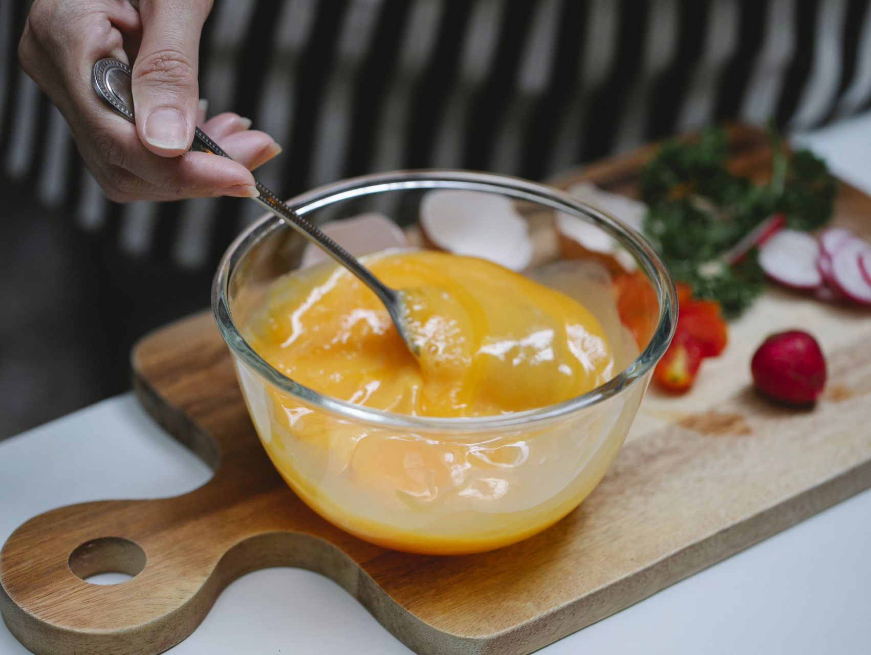 Person whisking yellow egg mixture in a glass bowl on a wooden cutting board with vegetables.