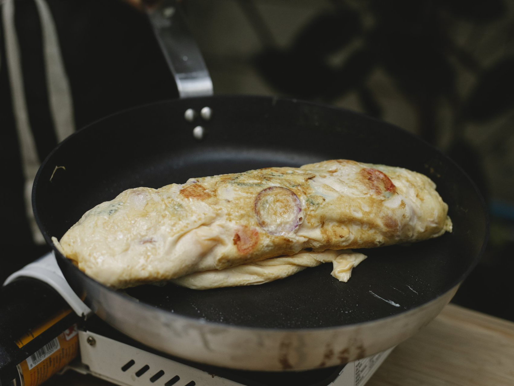 Omelet cooking in a pan on a portable burner; red tomatoes and onions visible.
