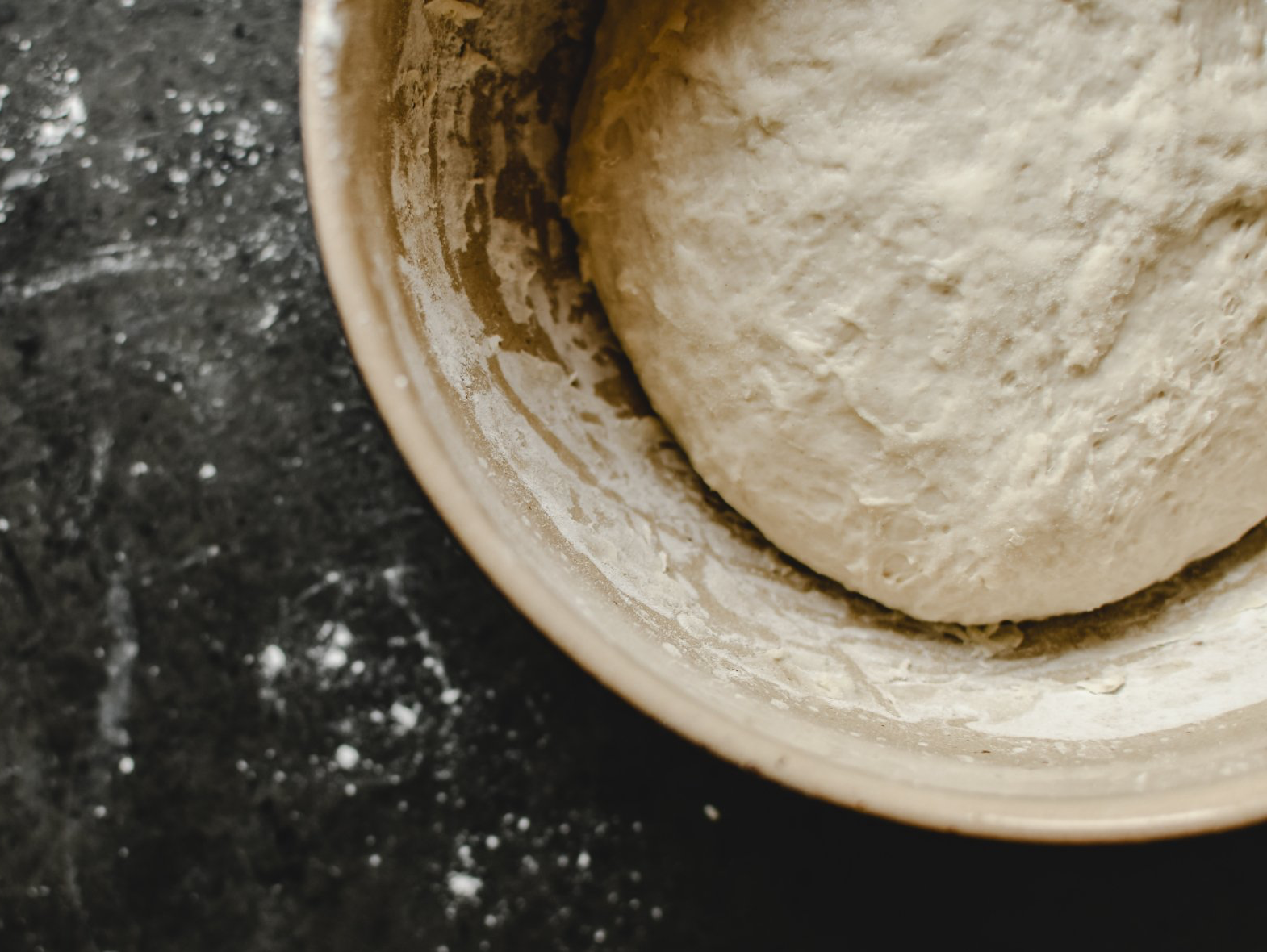 Dough ball in a ceramic bowl dusted with flour on a dark countertop.