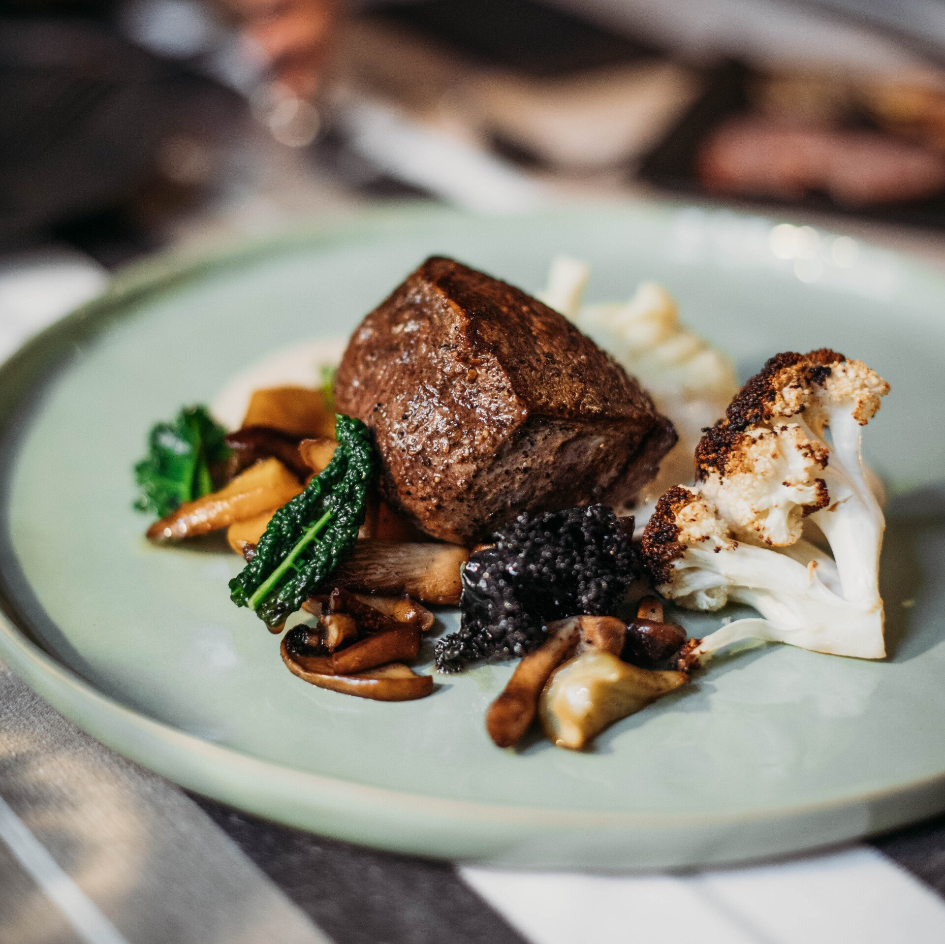 Plate of seared meat, cauliflower, mushrooms, and greens on a light green plate.