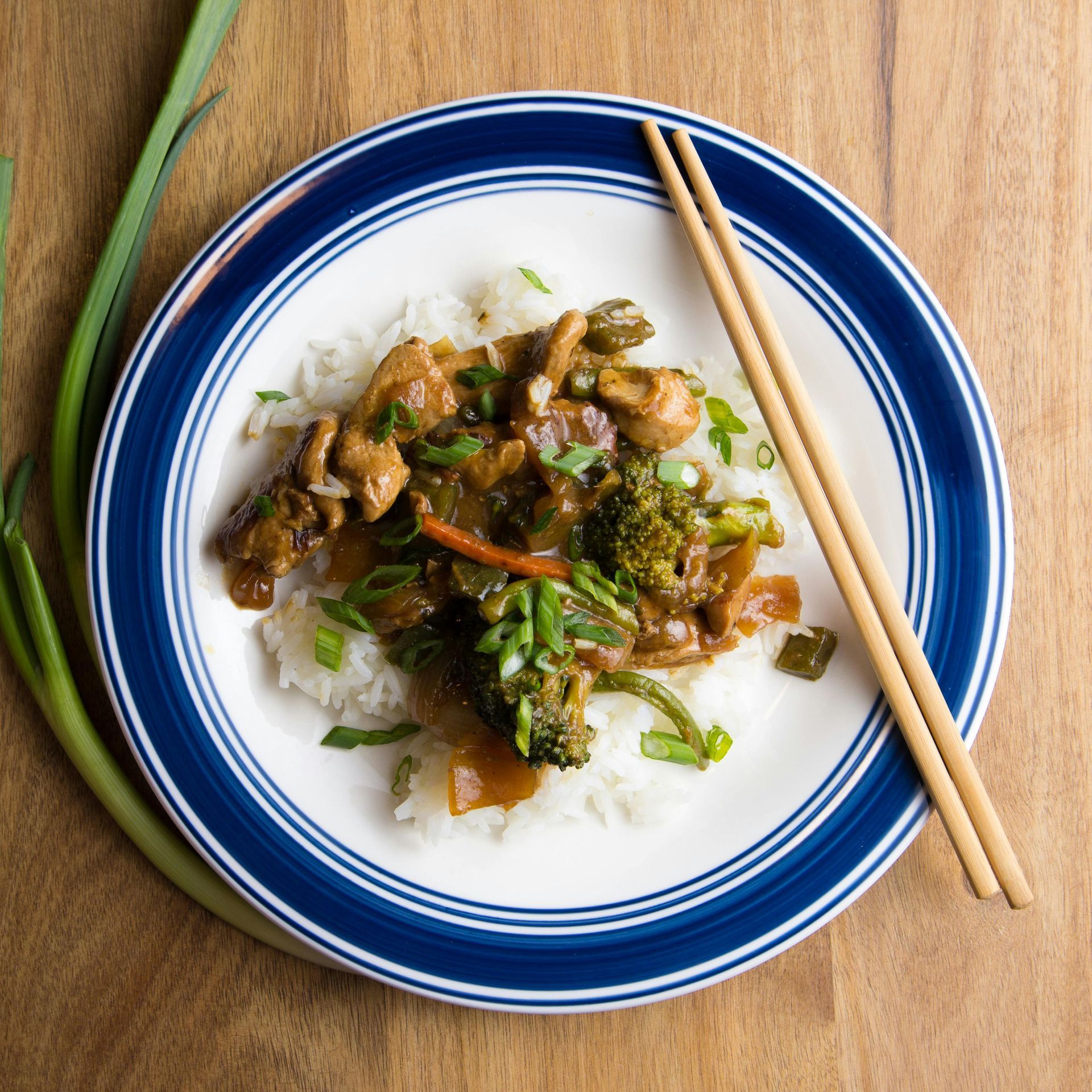 Chicken and broccoli stir-fry over rice, garnished with green onions, on a blue-rimmed plate with chopsticks.
