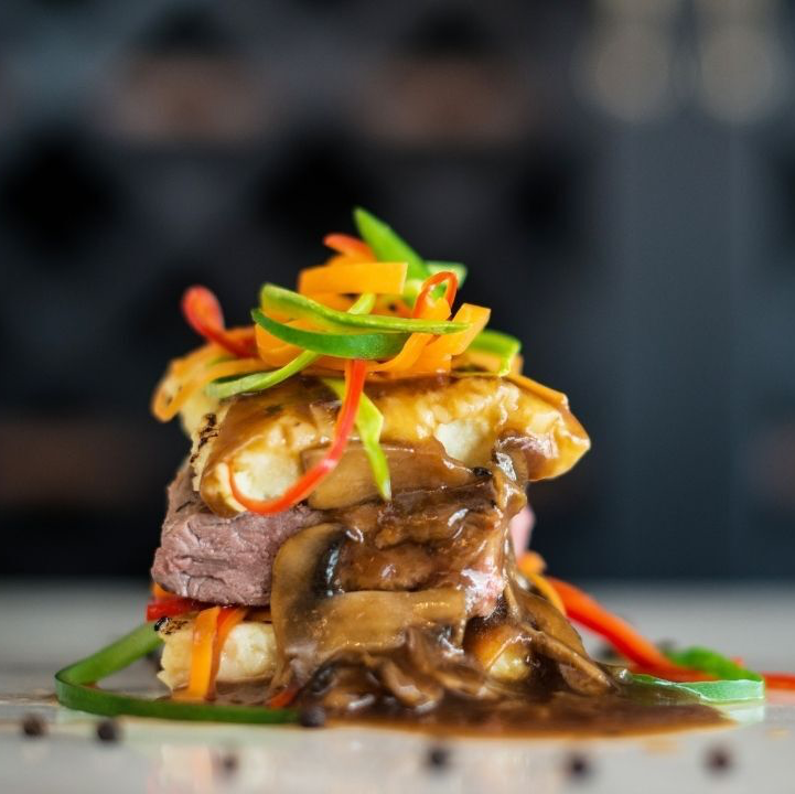 Stack of food with steak, mushrooms, gravy, and colorful vegetable garnish.