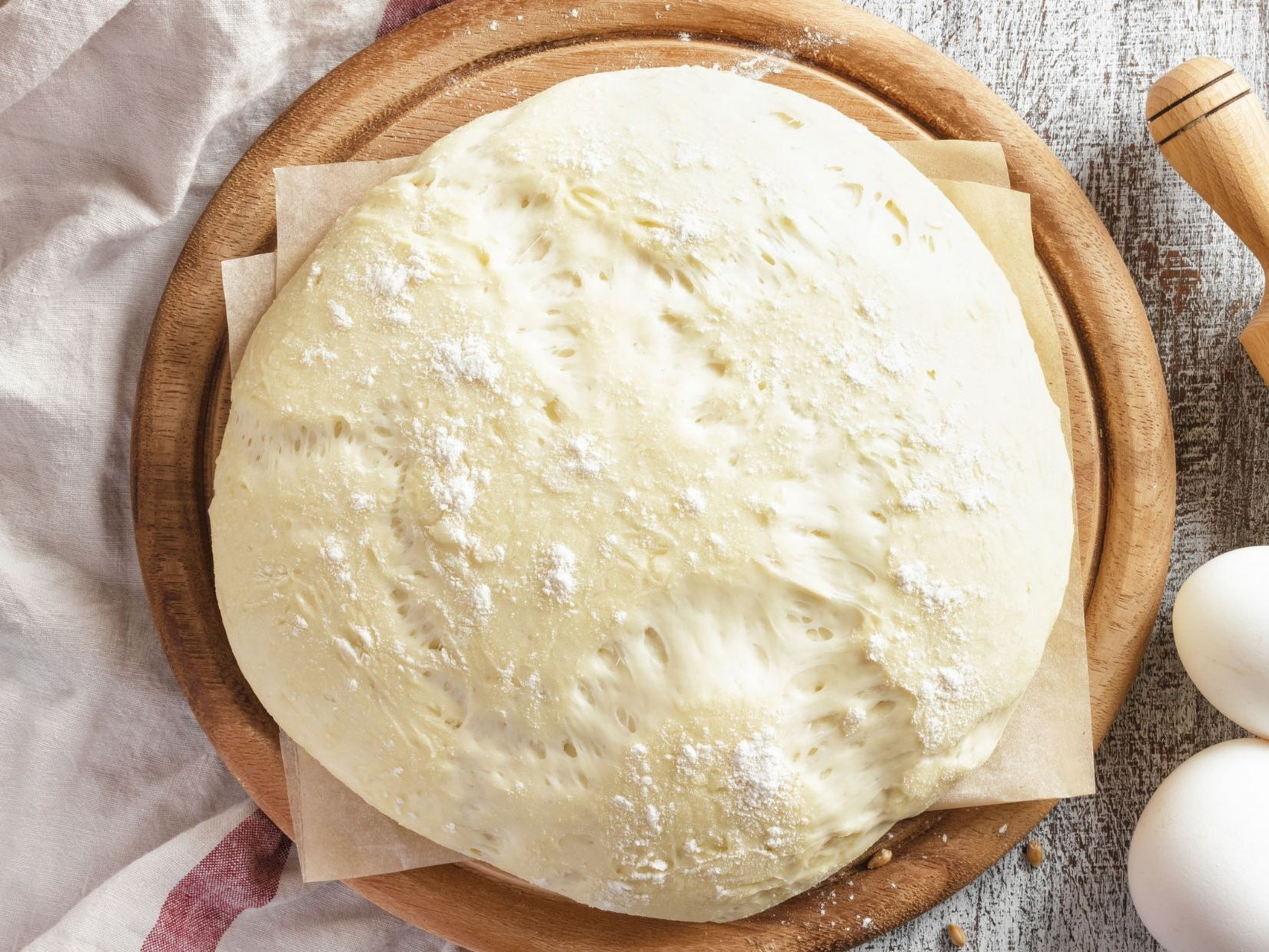 Dough on parchment paper, on a wooden board. Flour dusted, light-colored. Eggs and rolling pin in the background.