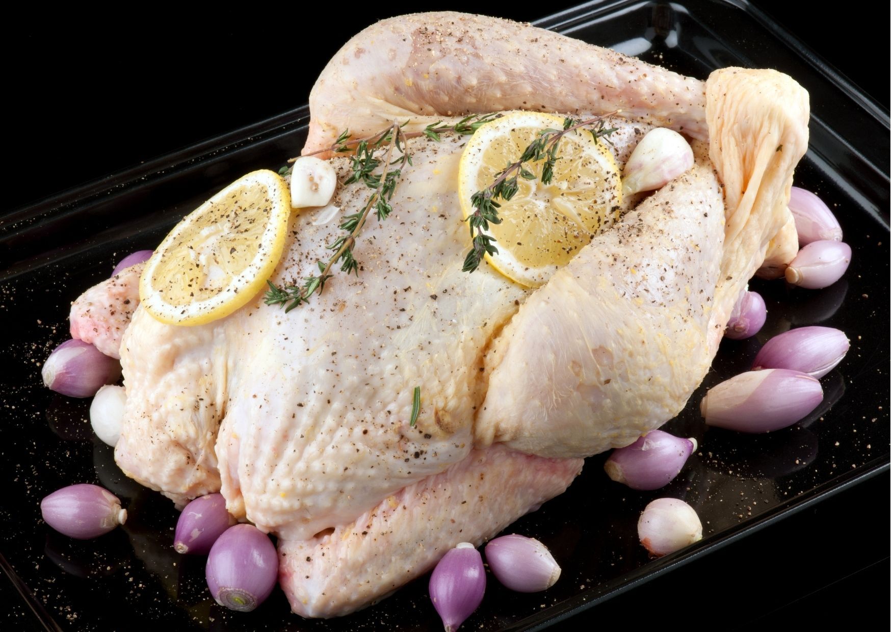 Raw chicken seasoned with lemon slices, thyme, garlic, and shallots on a black tray.